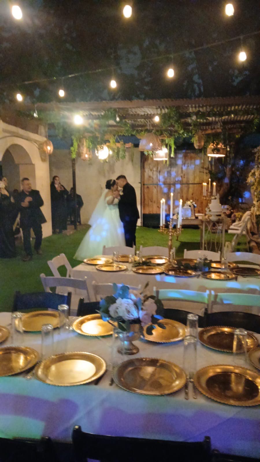 Novios bailando bajo luces de cadena en un jardín de eventos, con las mesas servidas y los invitados alrededor
